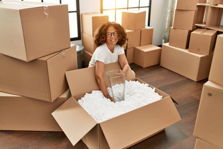 A woman surrounded by moving boxes is placing a glass into a box full of packing peanuts in the concept of 'What to Do with Packing Materials After Moving House to Slough'.